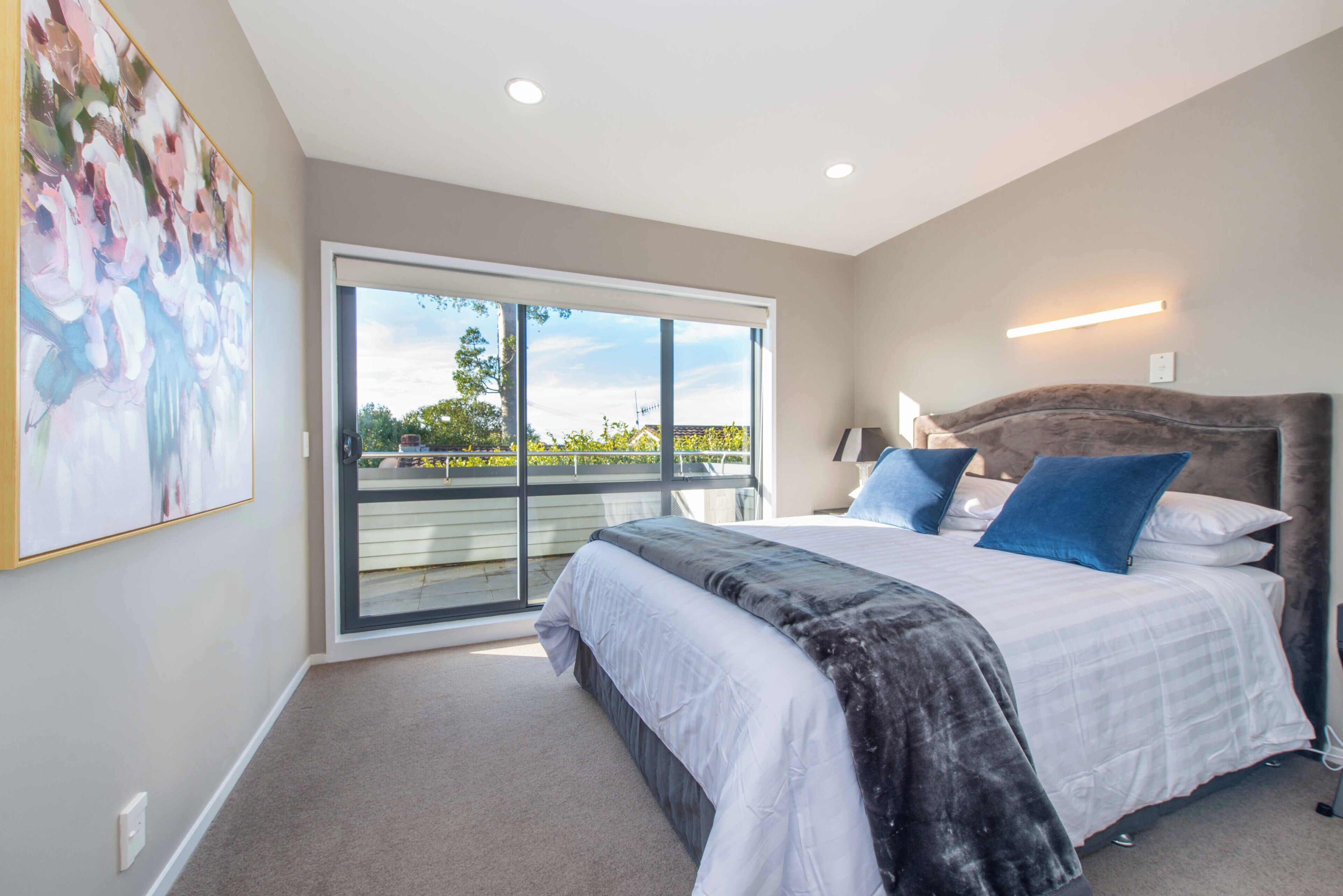 A bedroom with a large bed, a nightstand, a lamp, and a view of the outdoors through a sliding door.