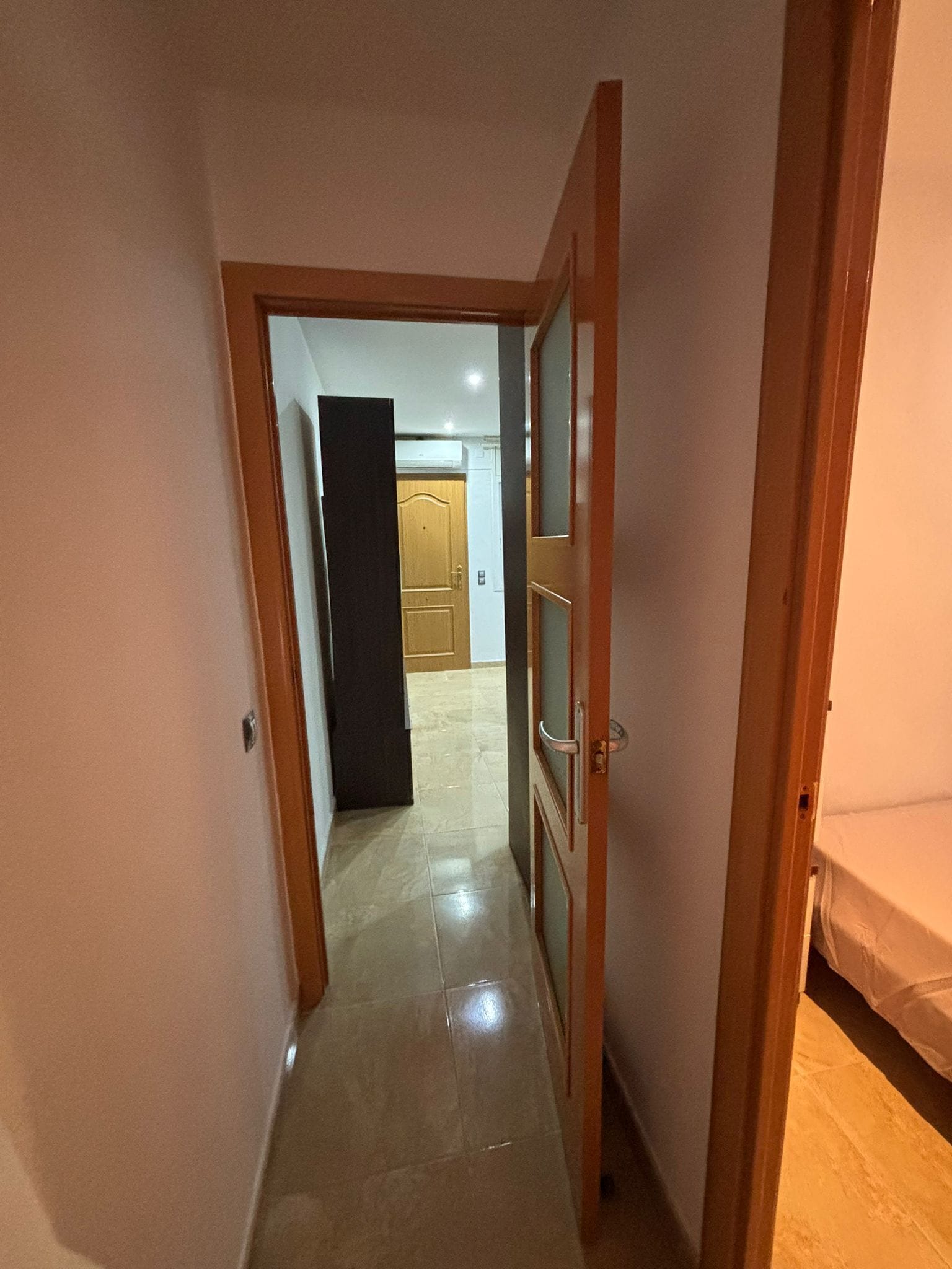 Un pasillo estrecho con una puerta de madera a la derecha, que da a una habitación con una cama.