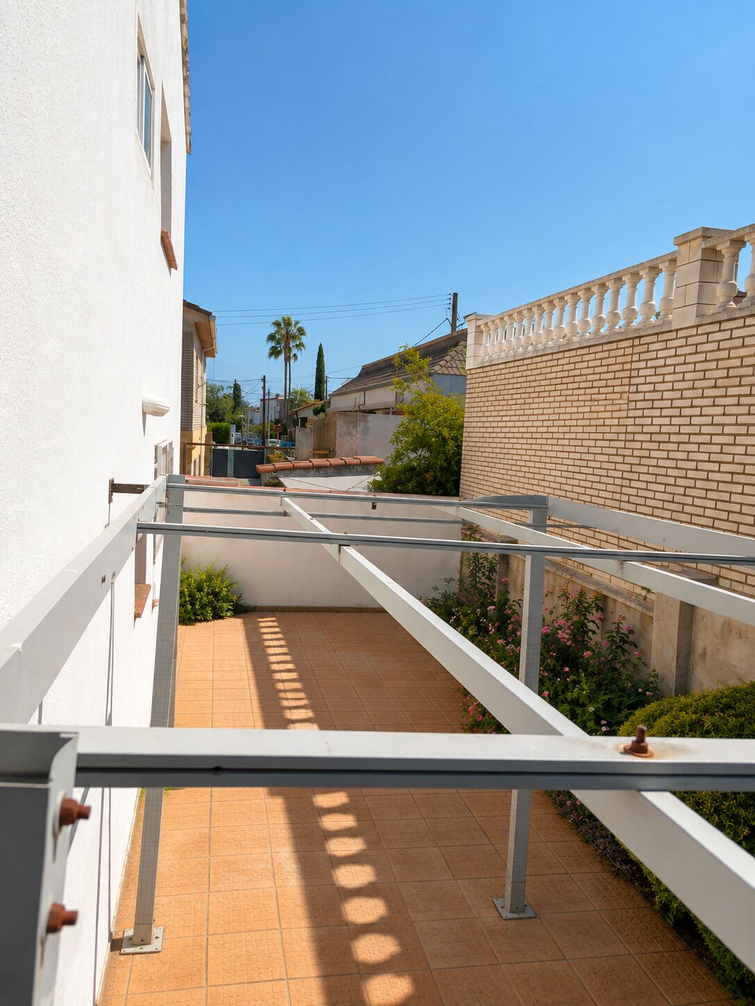 Un estrecho pasillo exterior con suelo de baldosas, barandillas blancas y una pared de ladrillo a un lado.