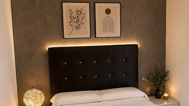 A bedroom with a bed, a nightstand with a lamp, a wall-mounted light fixture, and two framed artworks.