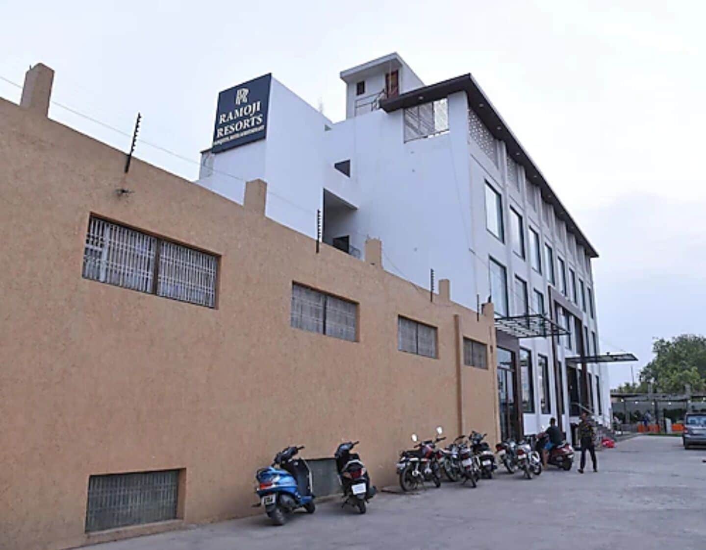 A building with a sign for Ramji Resorts, a parking area with multiple motorcycles, and a person standing near the entrance.