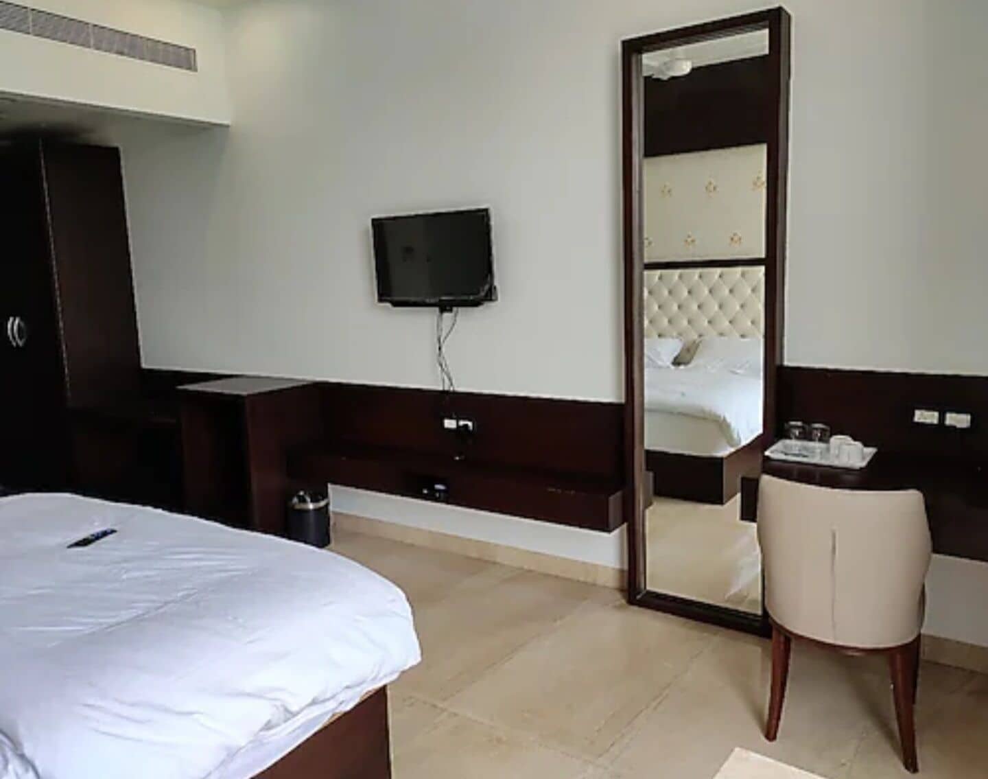 A hotel room with a bed, a TV mounted on the wall, a large mirror, a bedside table, and a chair.