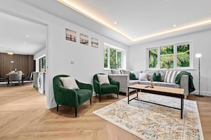 Ein modernes Wohnzimmer mit grünen Sesseln, einem grauen Sofa und einem Couchtisch. Große Fenster bieten Blick auf Grünflächen.