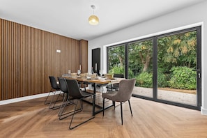 Ein moderner Essbereich mit einer Wandpaneele aus Holz, einem Esstisch für vier Personen und großen Fenstern mit Blick auf Grünflächen.
