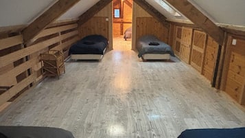 Um pequeno quarto no sótão com teto inclinado, paredes de madeira e uma porta central que dá para outro cômodo. Há duas camas encostadas nas paredes.