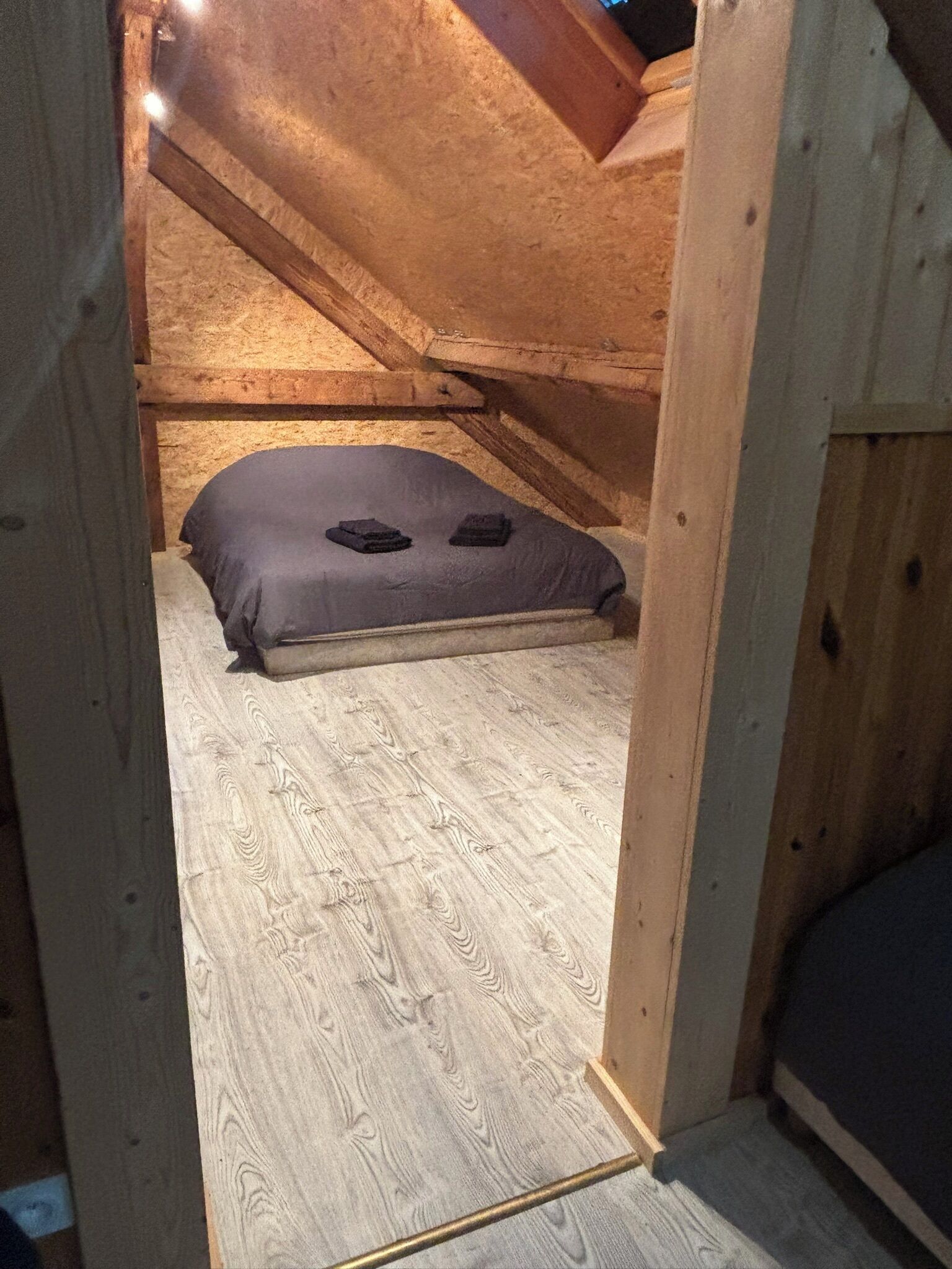 Quarto de sótão pequeno, com teto inclinado, uma cama de solteiro e chão de madeira.