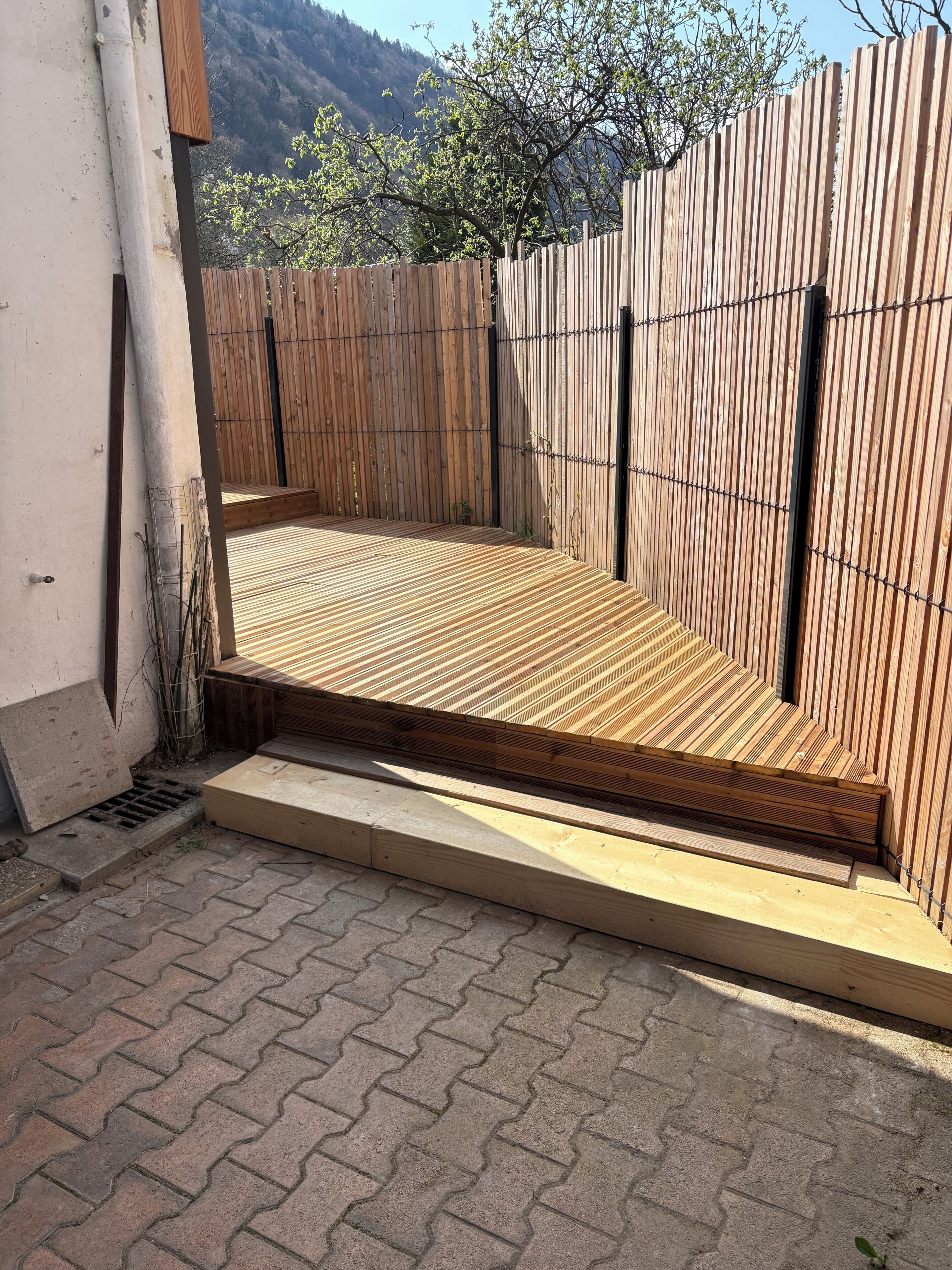 A wooden deck with a built-in bench, surrounded by a wooden fence, and a paved area with a drainpipe.