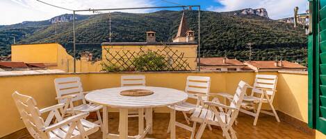 A terrace with a round table and chairs, a view of a mountain, and a building with a pointed roof.