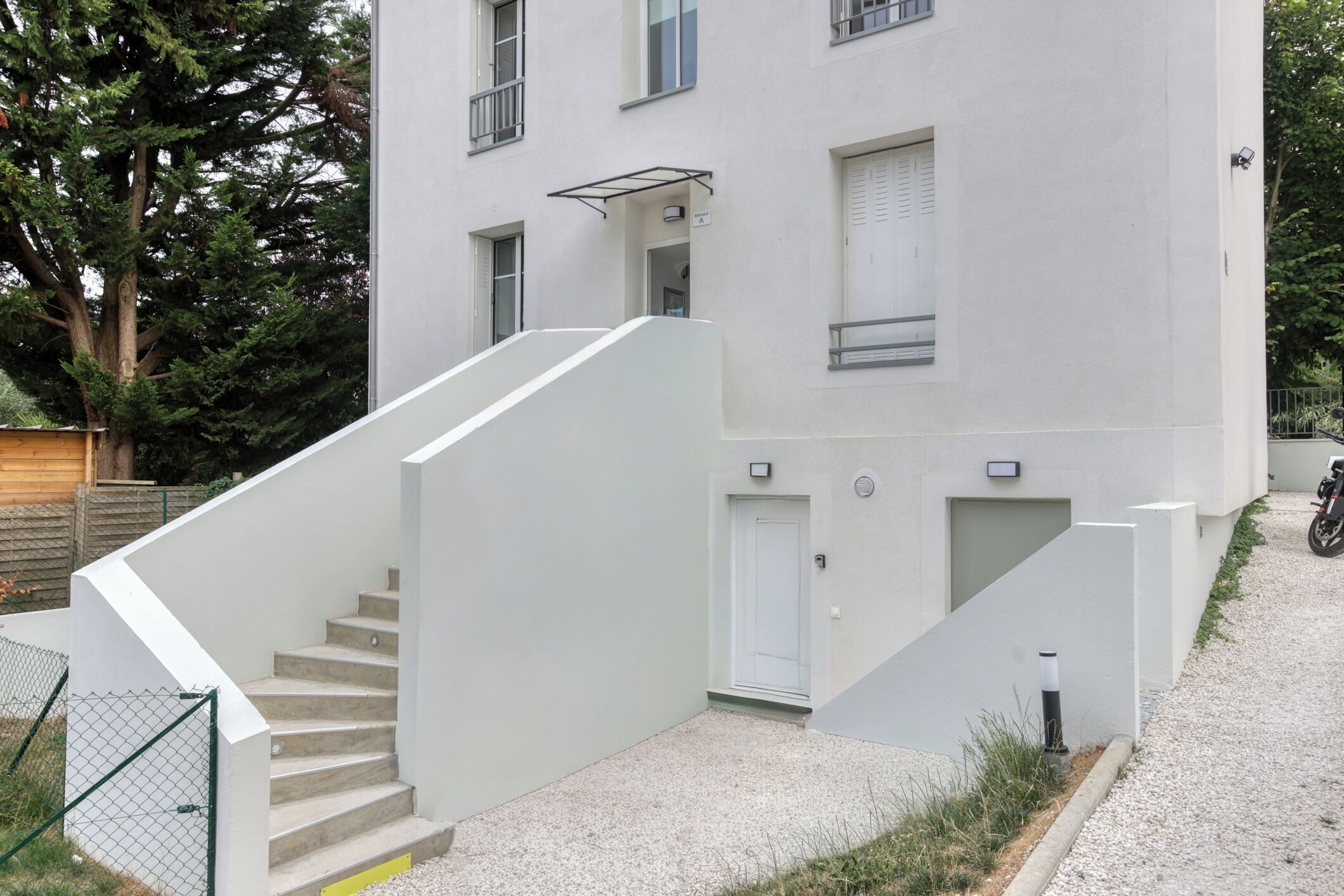 A modern two-story building with a staircase leading to the entrance, a covered outdoor area, and a vehicle parked nearby.