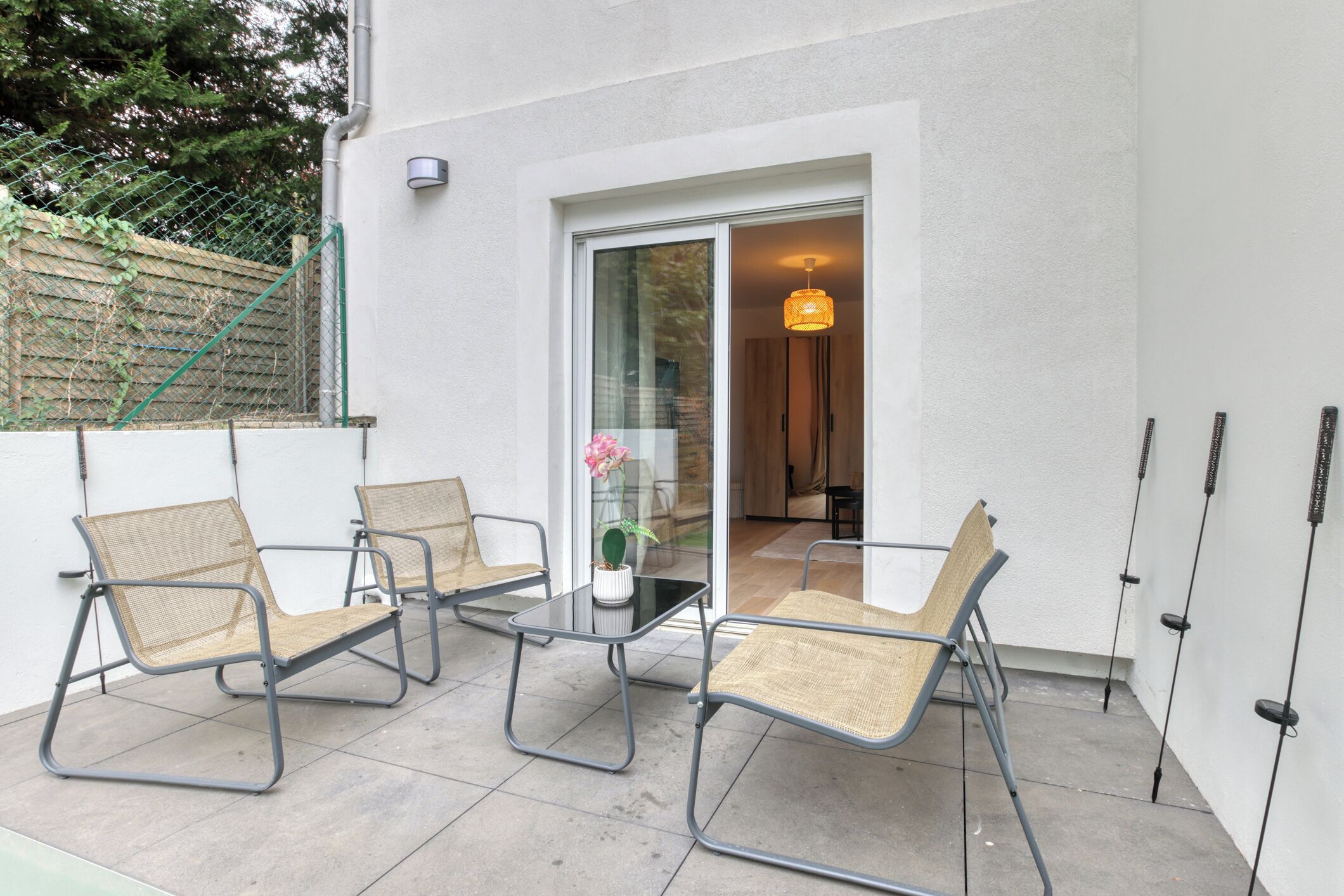 An outdoor patio with a glass table and four chairs, a potted plant, and a hanging light fixture.