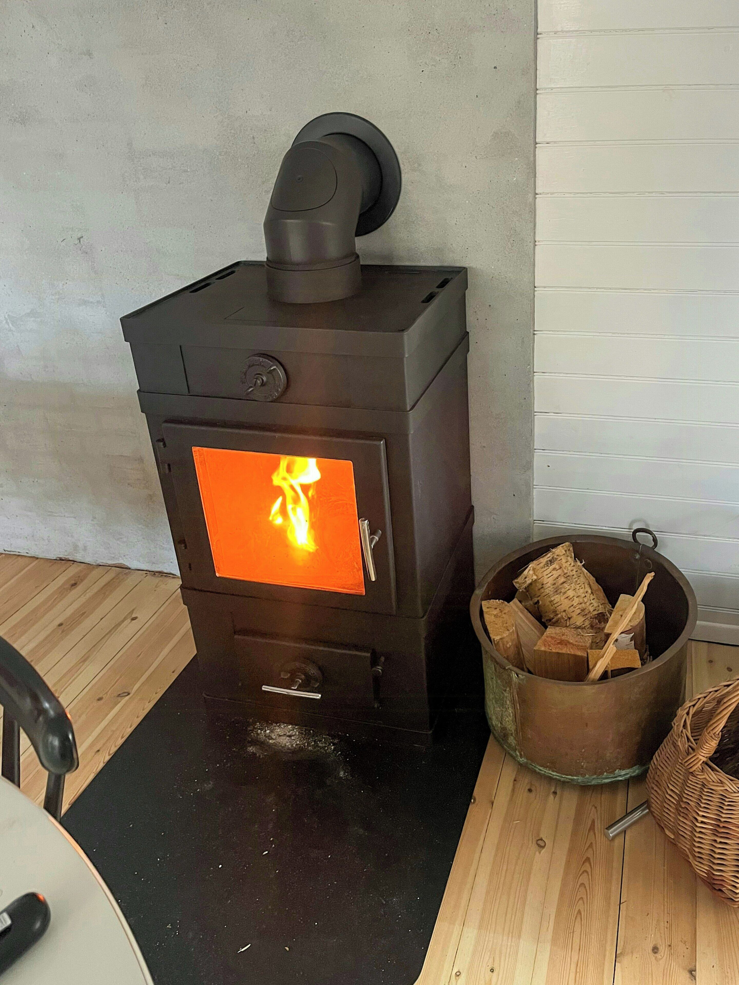 A wood-burning stove with visible flames, a firewood storage container, and a black mat on the floor.
