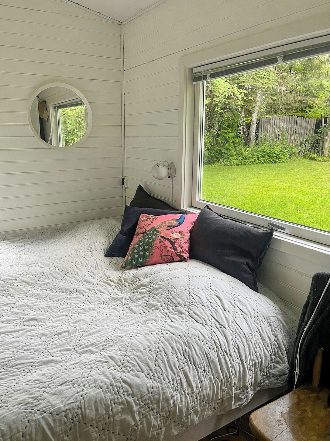 A bedroom with a bed, a round mirror, a window with a view of a green lawn, and a wall-mounted lamp.