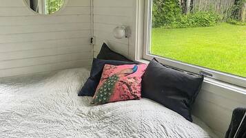 A bedroom with a bed, a round mirror, a window with a view of a green lawn, and a wall-mounted lamp.