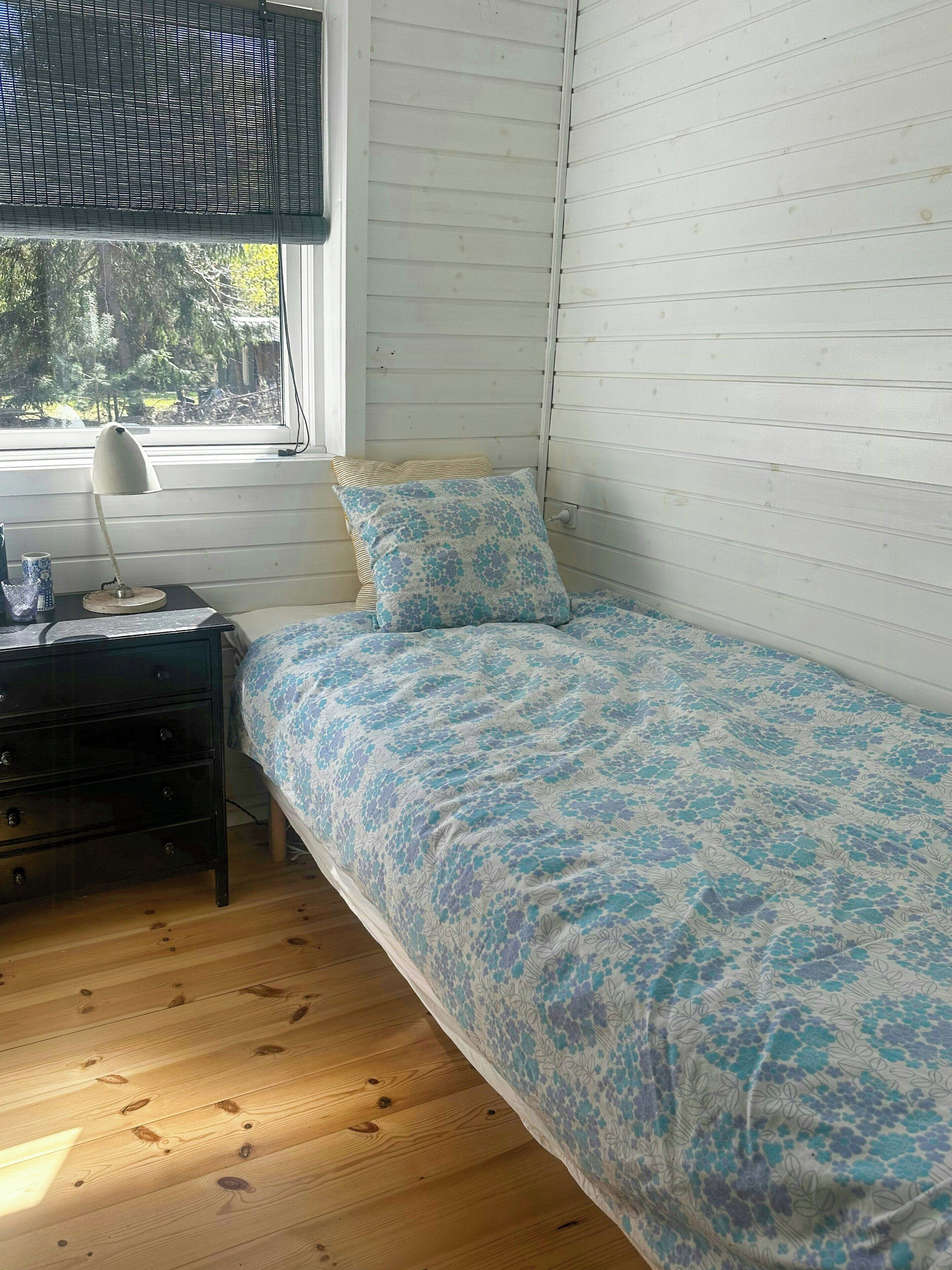 A bedroom with a single bed, a bedside table, a lamp, and a window with blinds.