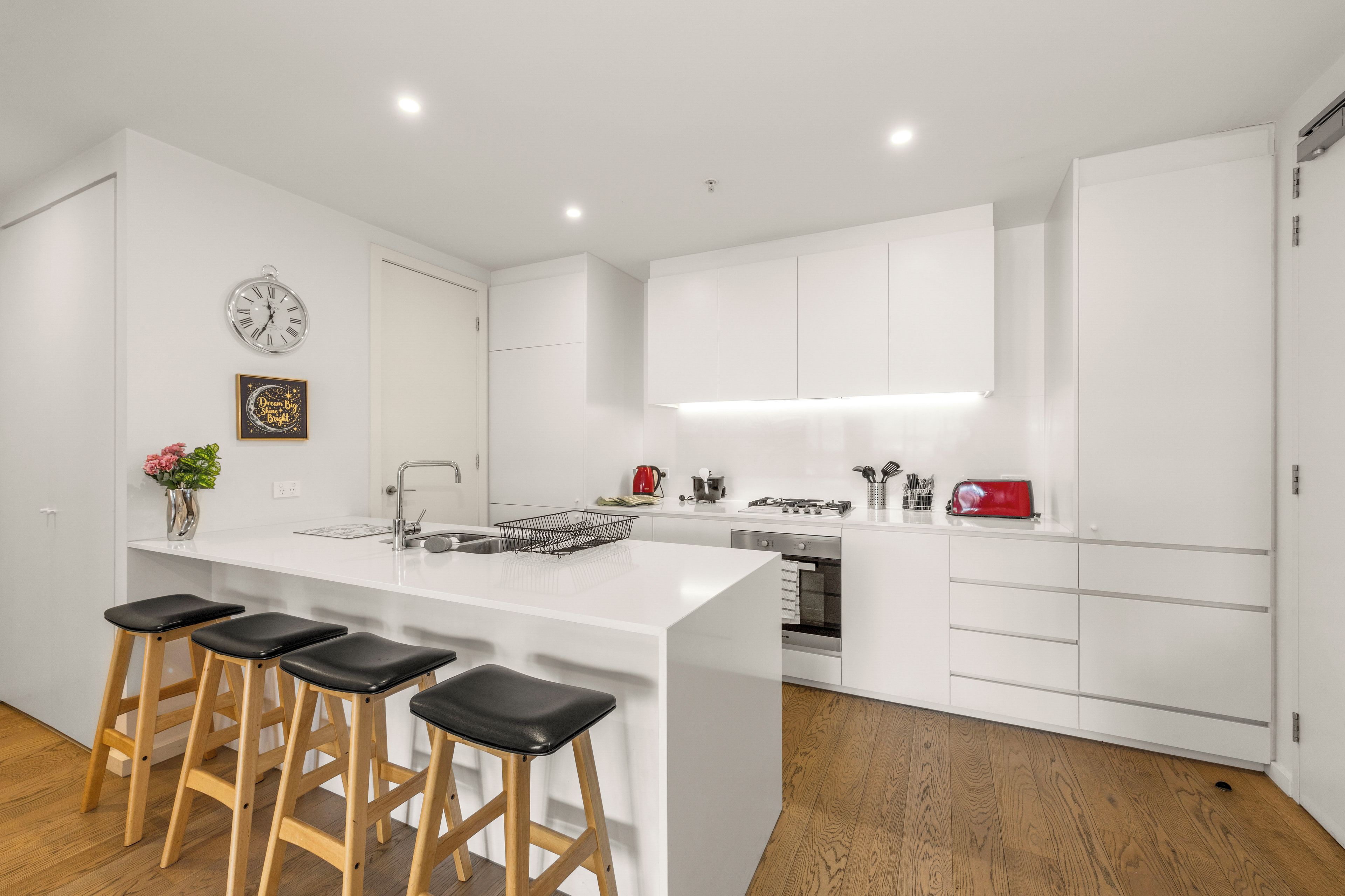 Une cuisine moderne avec des meubles blancs, un îlot central doté de tabourets, un grille-pain rouge et une horloge au mur.