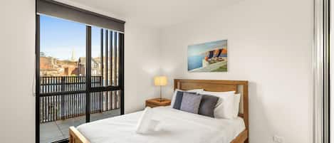 A hotel room with a large bed, a nightstand, a lamp, a painting, and a view of the city through a sliding door.
