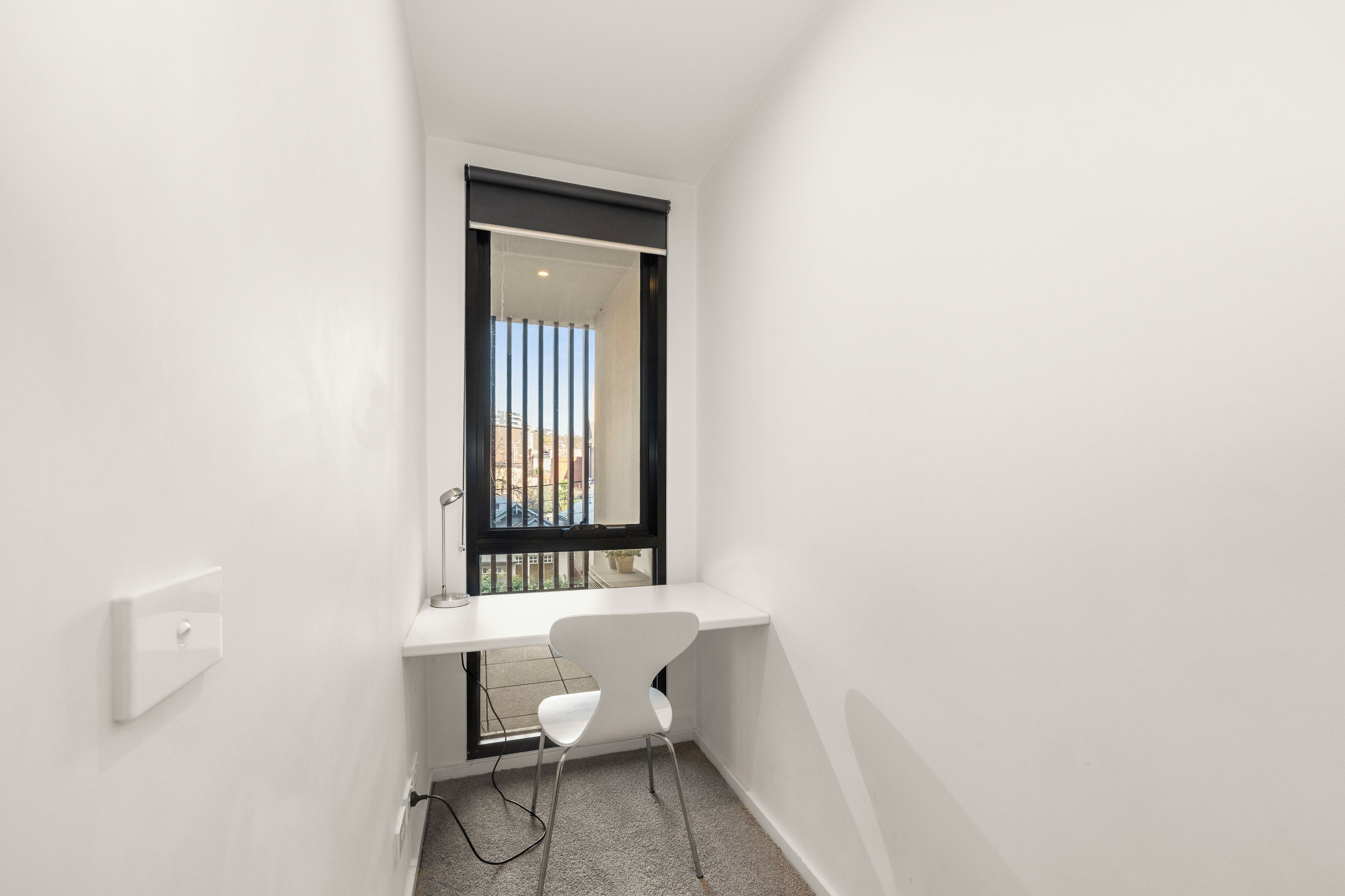 A small, narrow room with a white desk, a chair, and a window with a view of buildings.