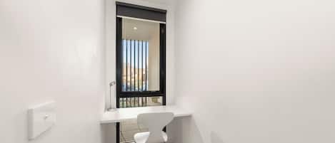 A small, narrow room with a white desk, a chair, and a window with a view of buildings.