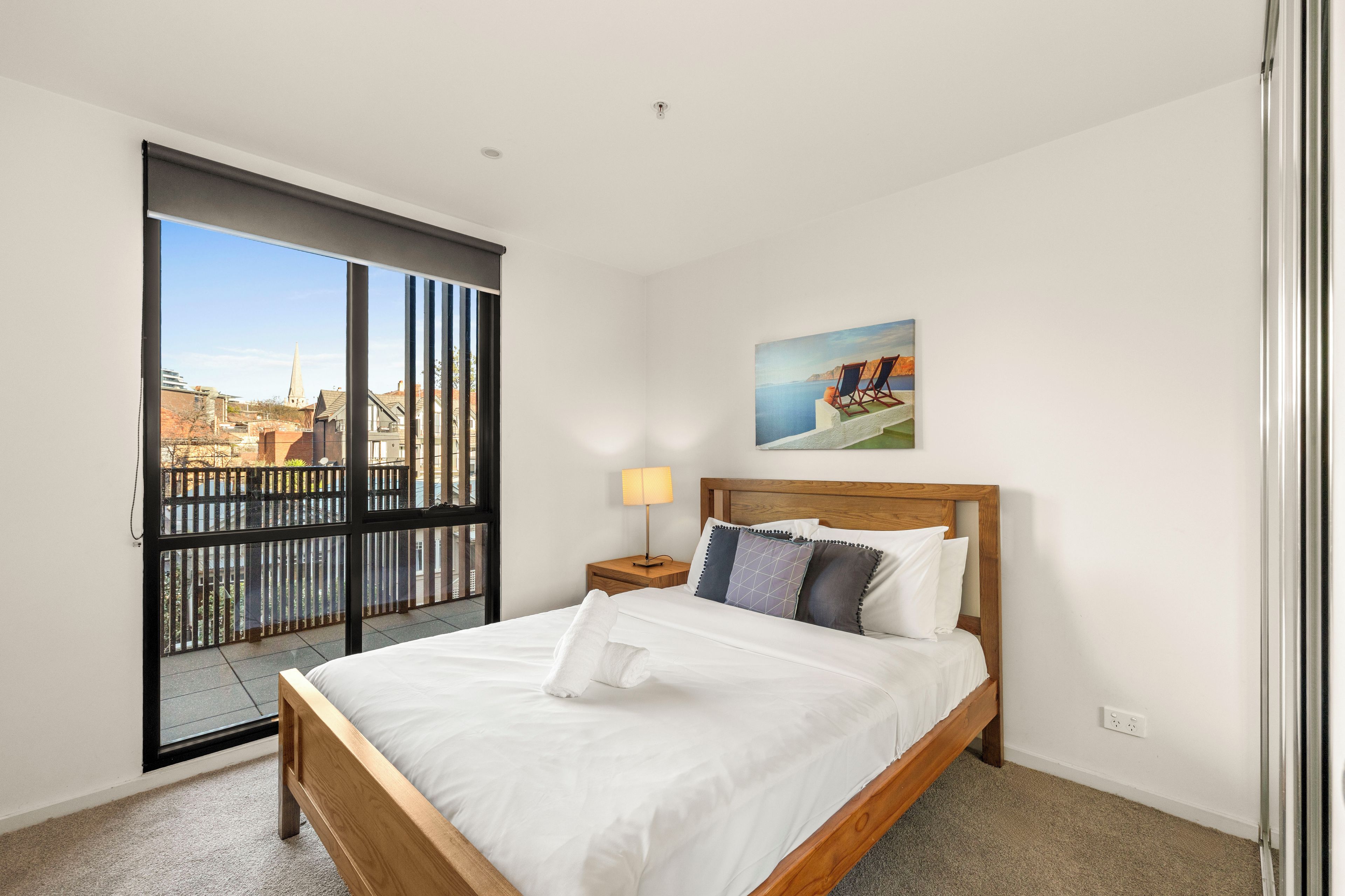 Una camera d'hotel con un letto grande, un comodino, una lampada, un quadro e vista sulla città attraverso una porta scorrevole.