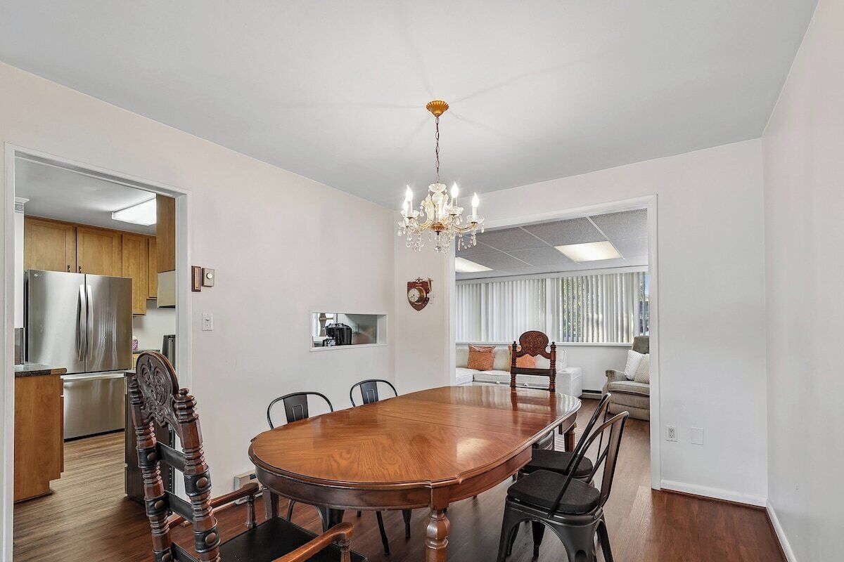 A dining area with a wooden table and chairs, a chandelier, and a kitchen in the background.