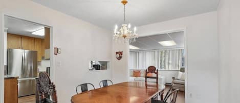 A dining area with a wooden table and chairs, a chandelier, and a kitchen in the background.