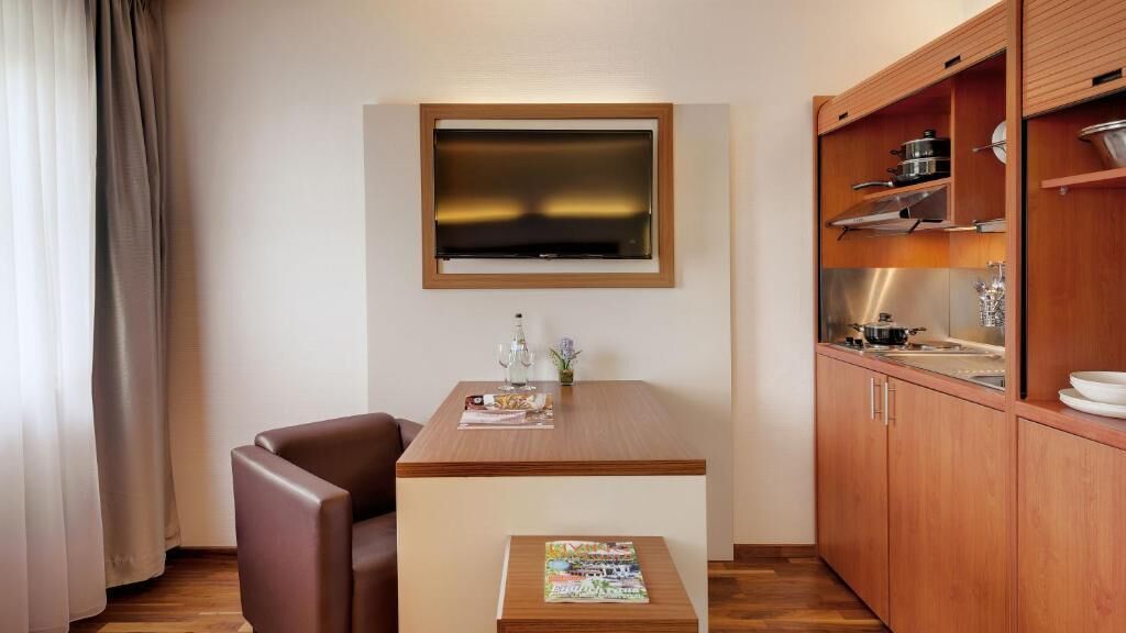 Ein kleines Hotelzimmer mit einem Holztisch, einem einzelnen braunen Stuhl, einem an der Wand befestigten Fernseher und einer Küchenzeile mit Holzschränken.