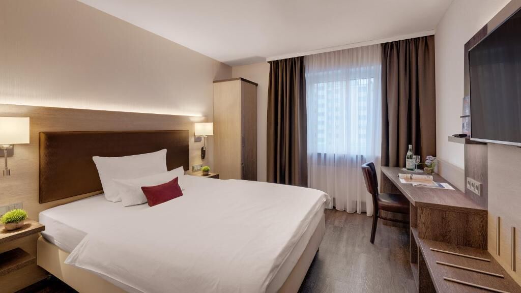 A hotel room with a large bed, a desk, a chair, a TV, and a window with curtains.