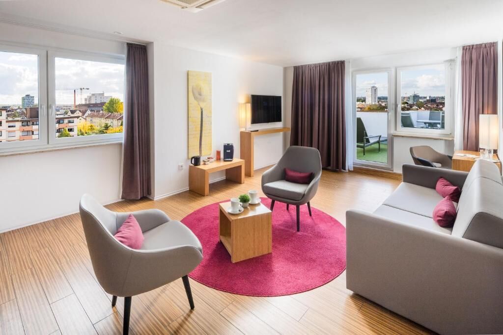 Ein modernes Wohnzimmer mit einer Couch, Sesseln, einem Couchtisch und einem Fernseher. Es gibt Fenster mit Vorhängen, ein Bild an der Wand und einen Blick auf Gebäude im Freien.