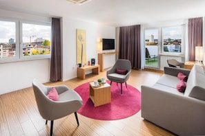 Een moderne woonkamer met een bank, stoelen, een salontafel en een televisie. Er zijn ramen met gordijnen, een schilderij aan de muur en een uitzicht op gebouwen.