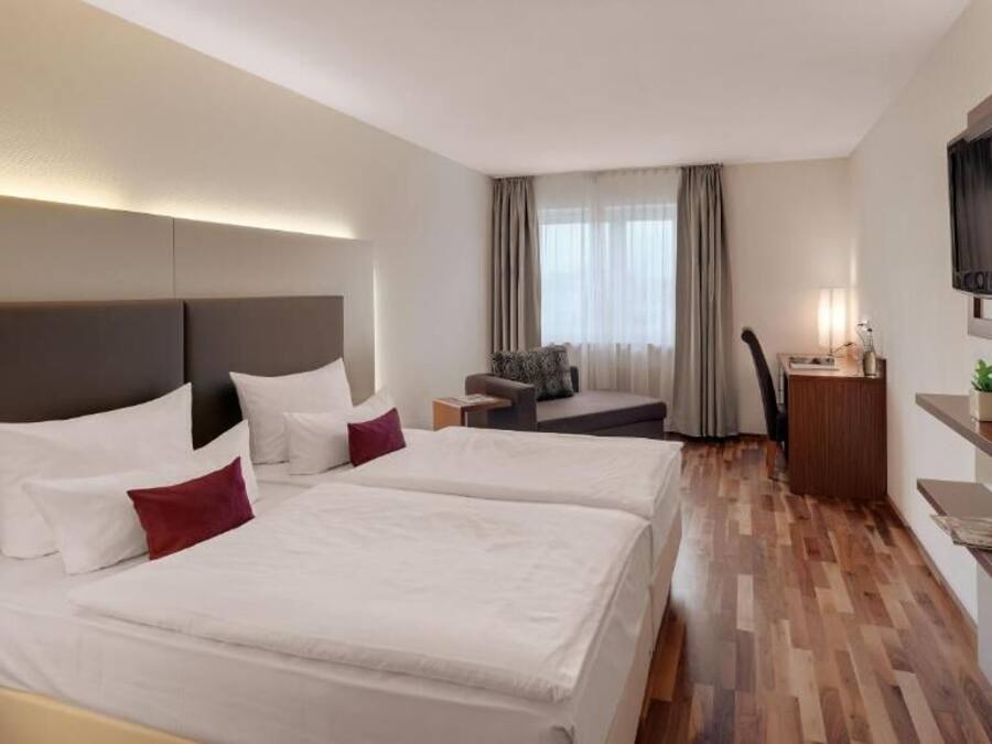 Ein Hotelzimmer mit zwei Betten, einem Schreibtisch, einem Stuhl, einem Fernseher und einem Fenster mit Vorhängen.