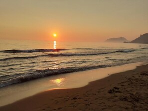 Eine Strandansicht bei Sonnenuntergang über dem Meer, sanfte Wellen umspülen das Ufer, und in der Ferne ist Land zu sehen.