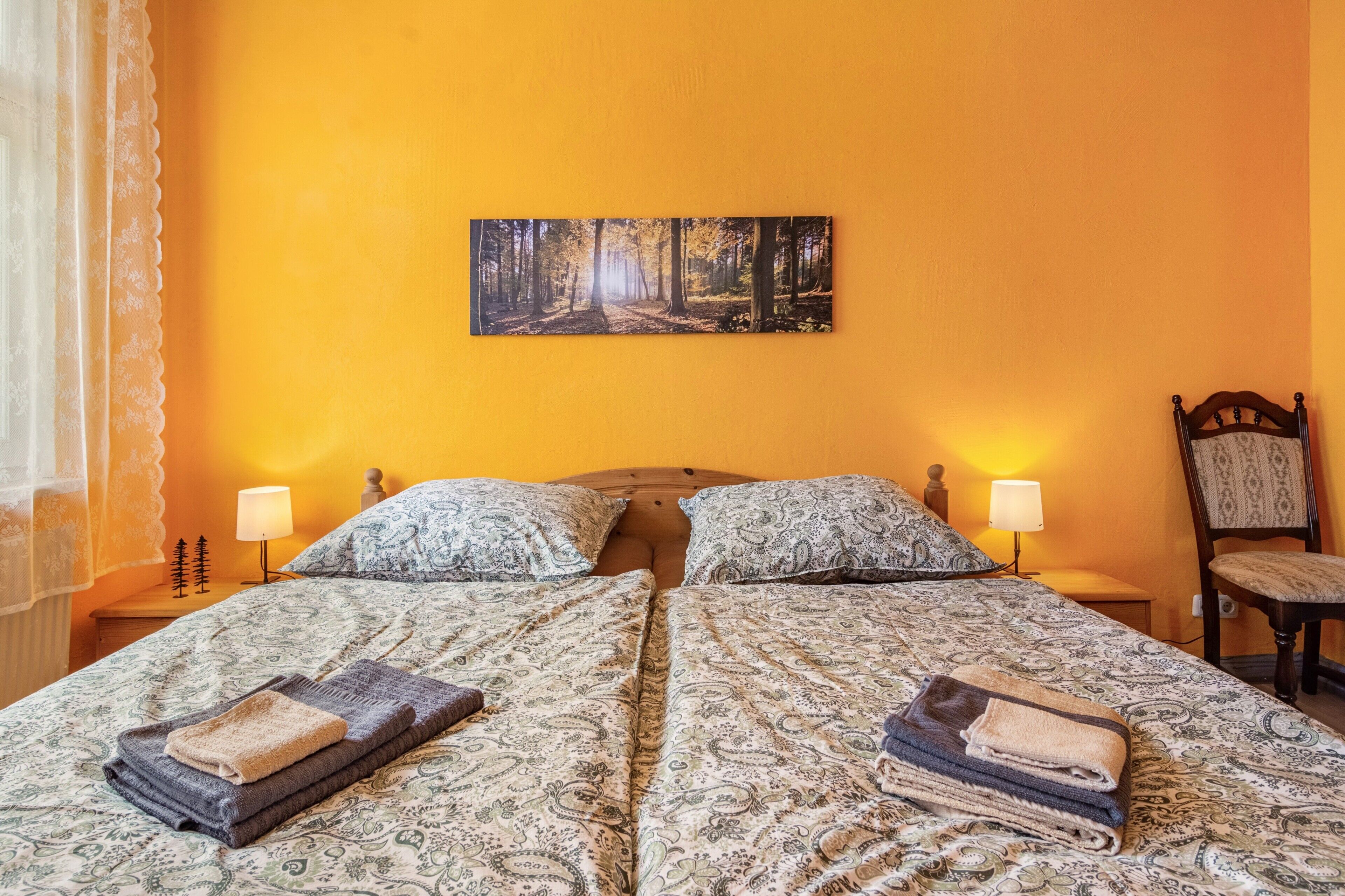 A double bed with patterned bedding and two pillows, a bedside table with a lamp, a chair with a patterned cushion, and a forest-themed painting on the wall.