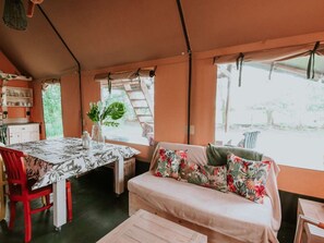 A tented accommodation with a dining table, red chairs, a sofa with floral cushions, and a view of greenery outside. - Kampari - Luxe Safaritent - De Jungle by Interhome (Delfstrahuizen)