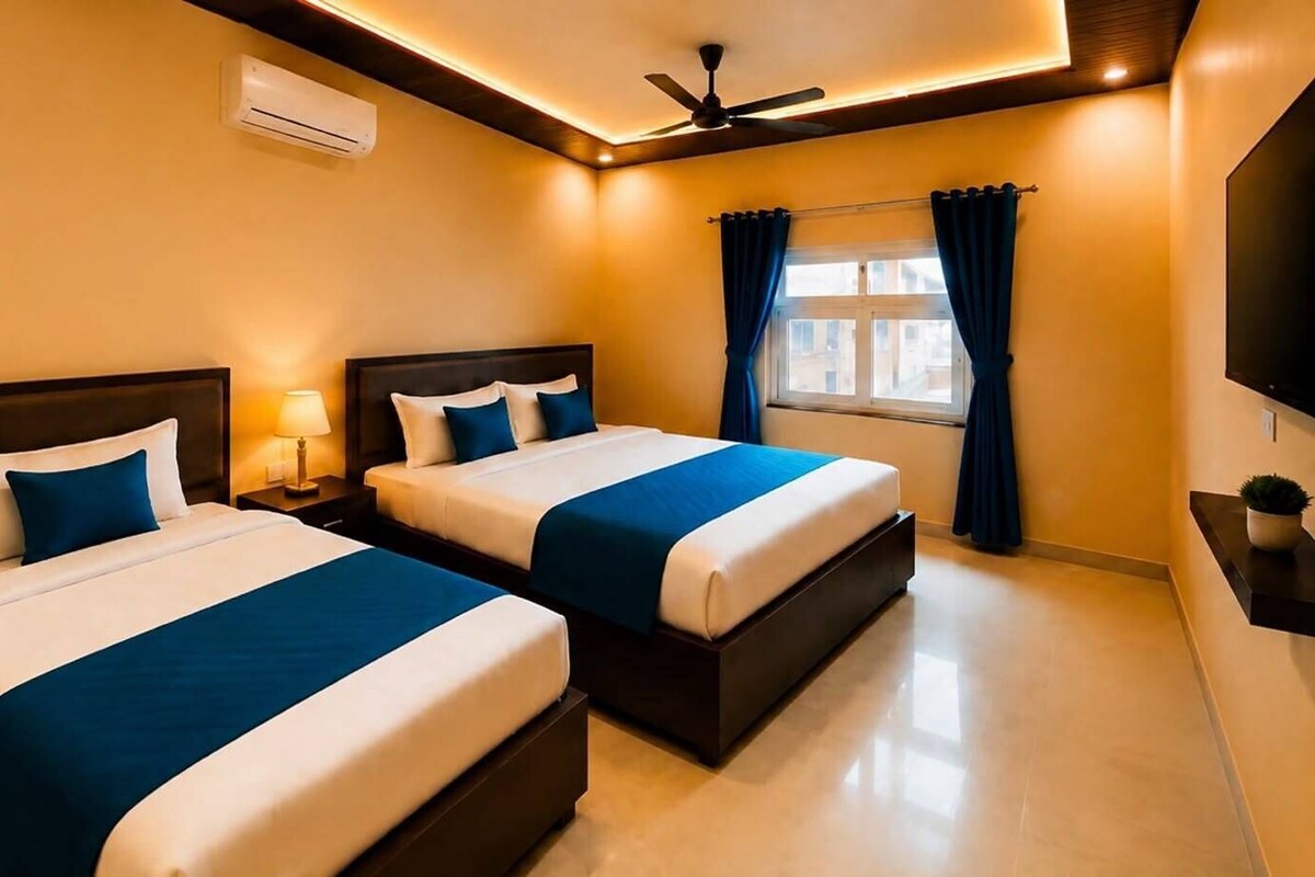 Een hotelkamer met twee bedden, een plafondventilator, een airconditioningapparaat, een raam met gordijnen en een flatscreen televisie die aan de muur is bevestigd.