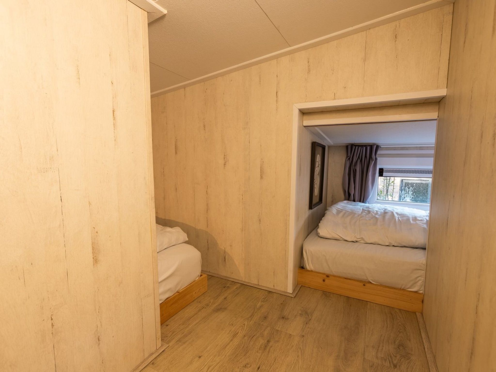 A small, single-room accommodation with a wooden floor and walls, a bed with white bedding, and a window with curtains.