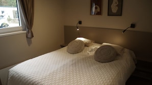 Uma cama com cobertura branca acolchoada, dois travesseiros decorativos, uma mesa de cabeceira com abajur e uma janela com cortinas.