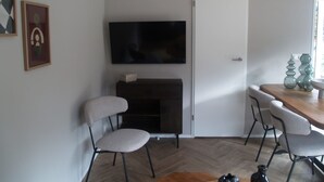 Um espaço de estar compacto com televisão de tela plana na parede, um armário de madeira escura, duas cadeiras, uma mesa de jantar com um vaso e um piso com padrão herringbone.