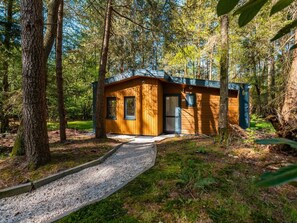 Een klein houten huisje met een grindpad dat naar de ingang leidt, omgeven door bomen.