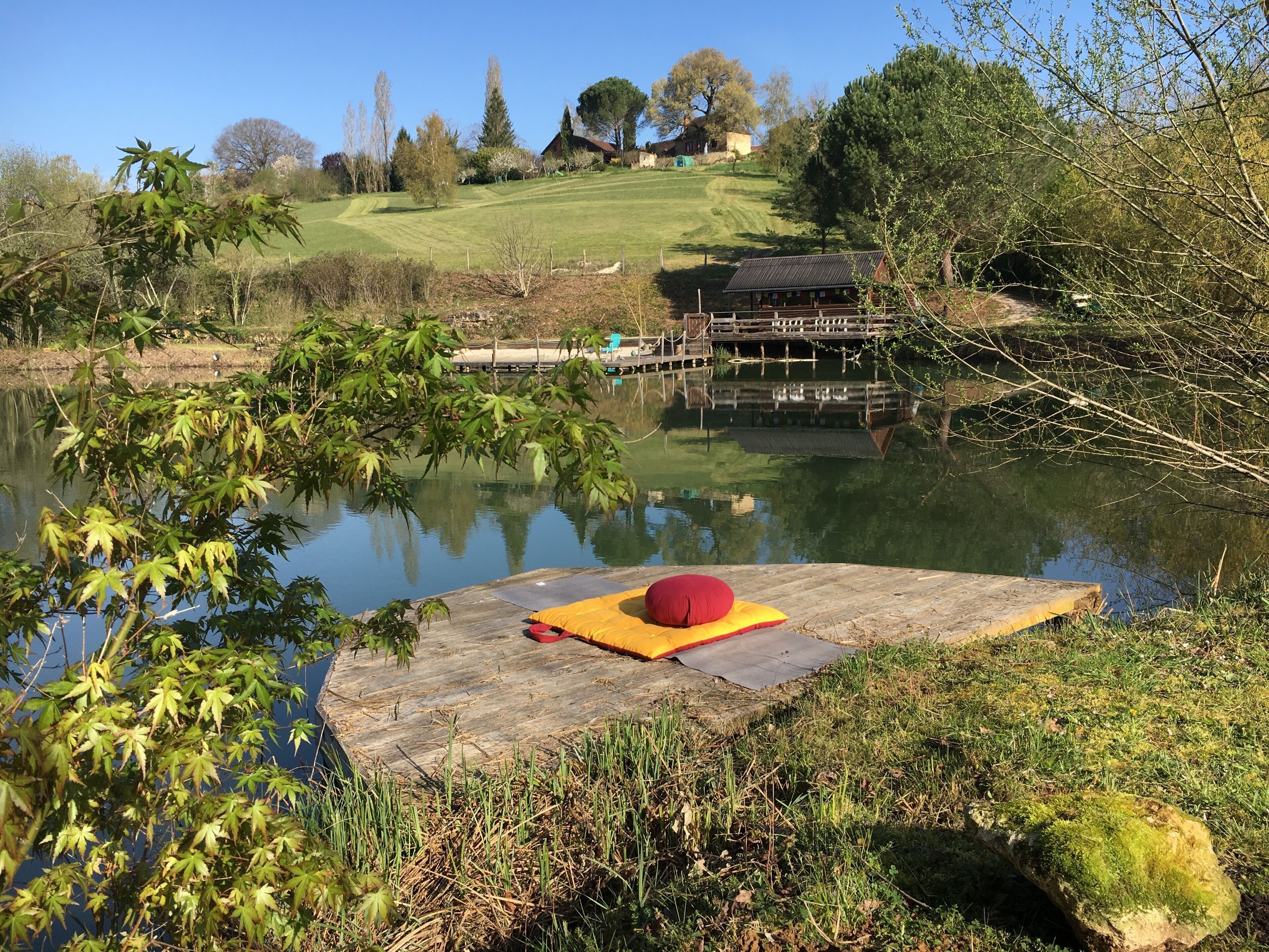 A wooden dock with a red cushion, a calm lake, and a grassy hill with trees in the background.