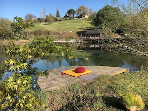 Ein Holzsteg mit einem roten Kissen, ein ruhiger See und ein grasbewachsener Hügel mit Bäumen im Hintergrund.