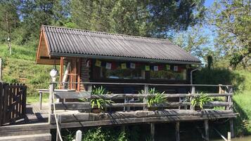 A wooden cabin with a metal roof, situated on stilts over water, surrounded by greenery.