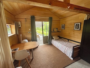A compact room with a bed, a table, and a chair. There is a window with curtains, a door leading outside, and a wall-mounted shelf with pictures.