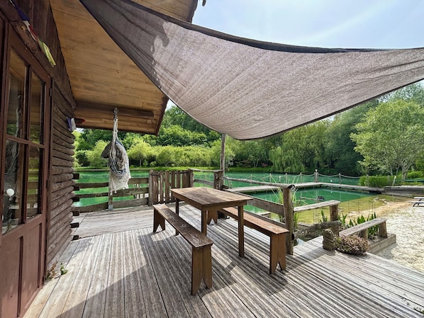 A wooden deck with a table and benches, a hanging chair, and a view of a pond and greenery.