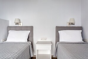 A hotel room with two beds, each with a grey headboard and white bedding. There are bedside tables with lamps and a small white nightstand between the beds. - Magical Sunsets & Stunning Panoramic Views (Tinos)