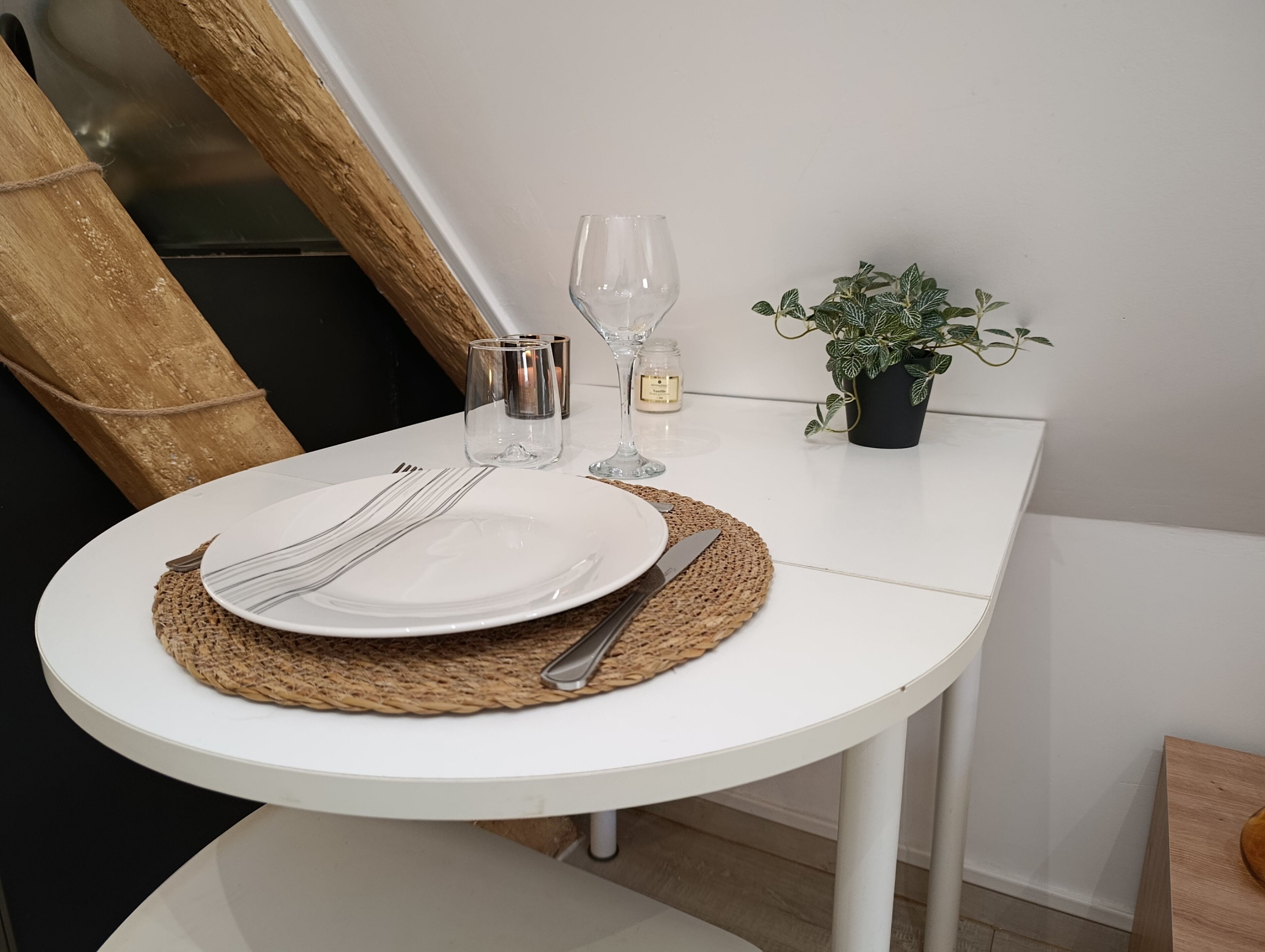 Une table ronde, recouverte d’une nappe blanche et agrémentée d’un chemin de table tissé, est mise pour le dîner : on y trouve une assiette blanche, des couverts, un verre et une bougie.