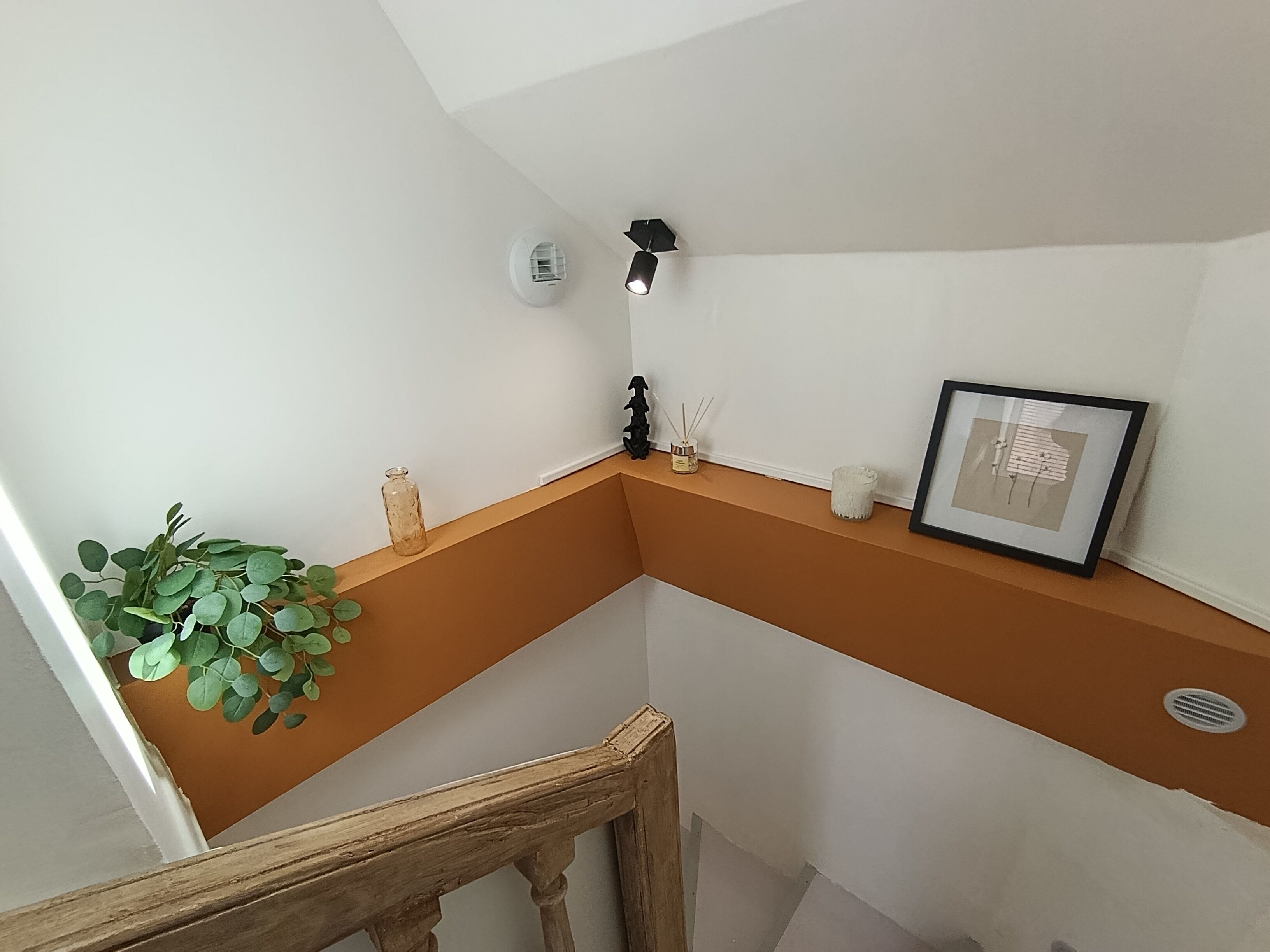 Een hoek van een kamer met een witte muur, een bruin plankje, een bloempot, een lijst met een foto en een rookmelder.