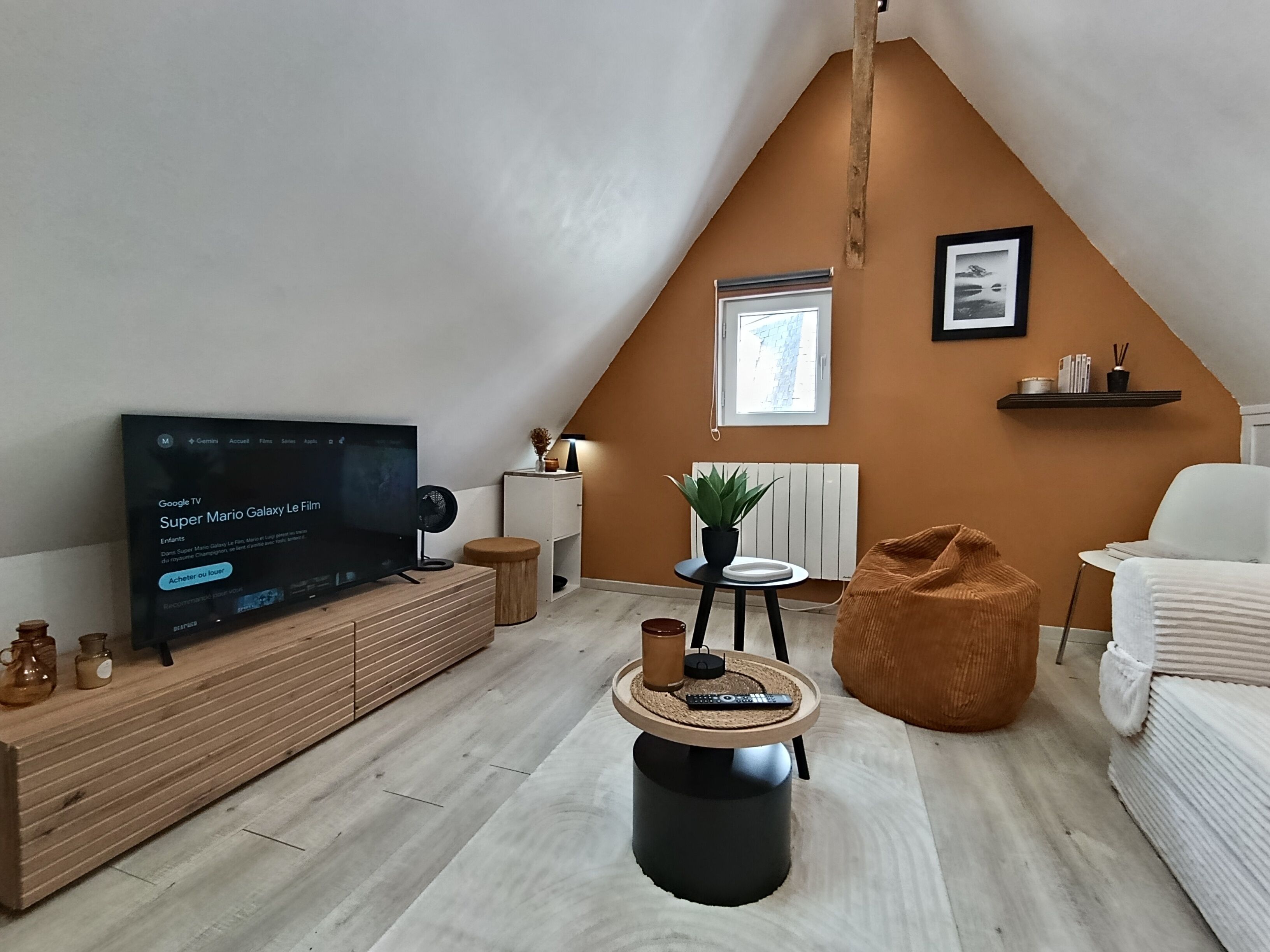 Een moderne woonkamer met een schuin dak, een flatscreen televisie, een ronde salontafel, een relaxfauteuil en een witte bank.