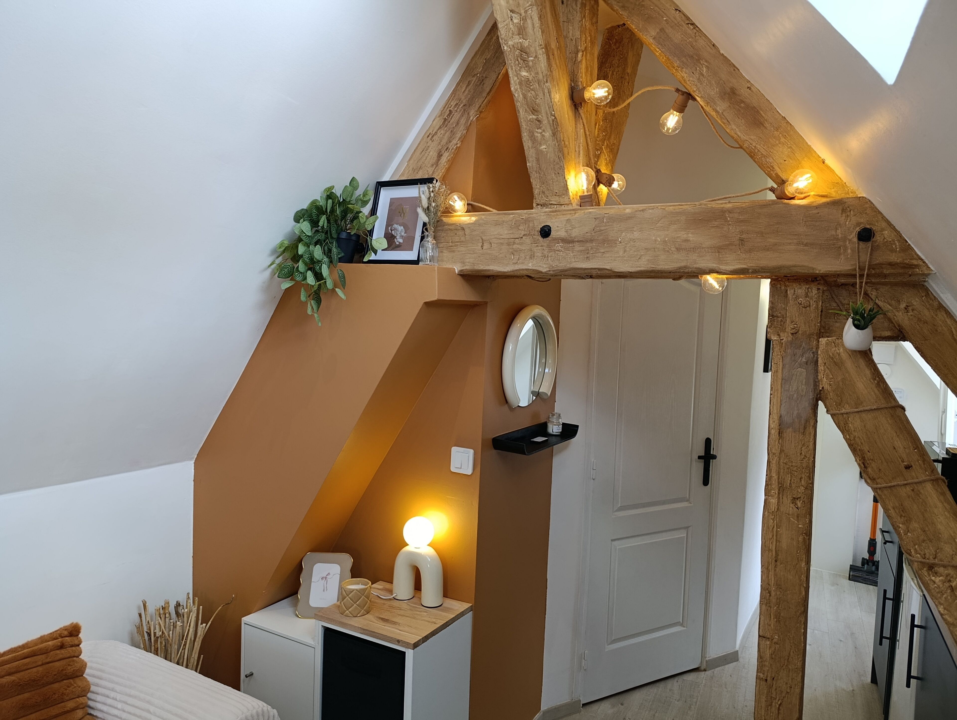 Une pièce avec un plafond en pente, des poutres en bois, une porte blanche, une table de chevet avec une lampe et une petite plante.