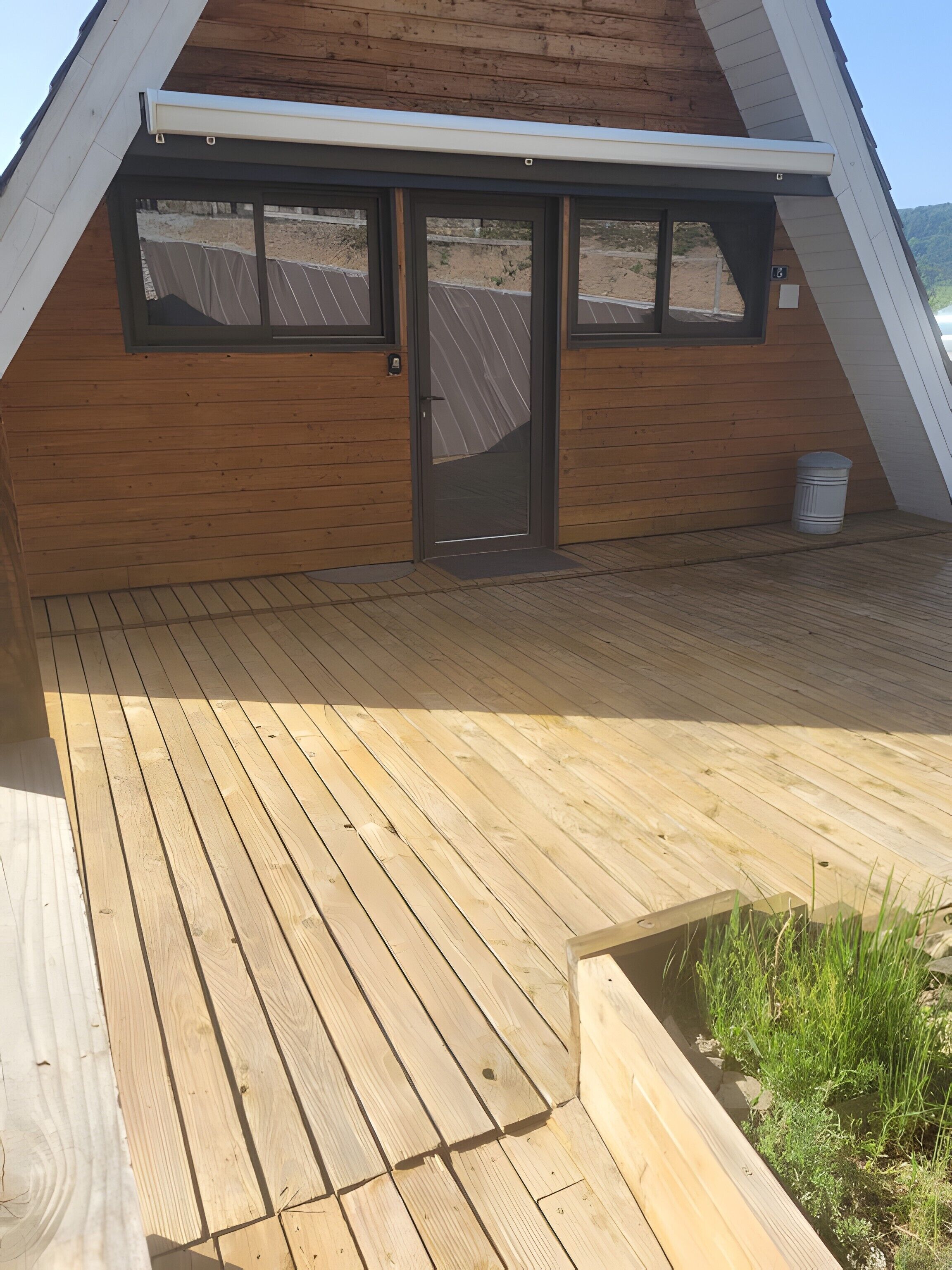 A wooden deck with a door and windows, a small trash bin, and a grassy area.