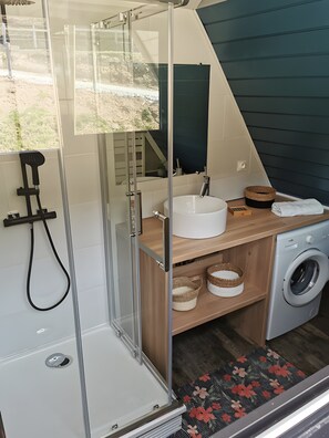 Bany compacte amb dutxa de vidre, un lavabo, un mirall, una rentadora i una petita estant amb cistelles.